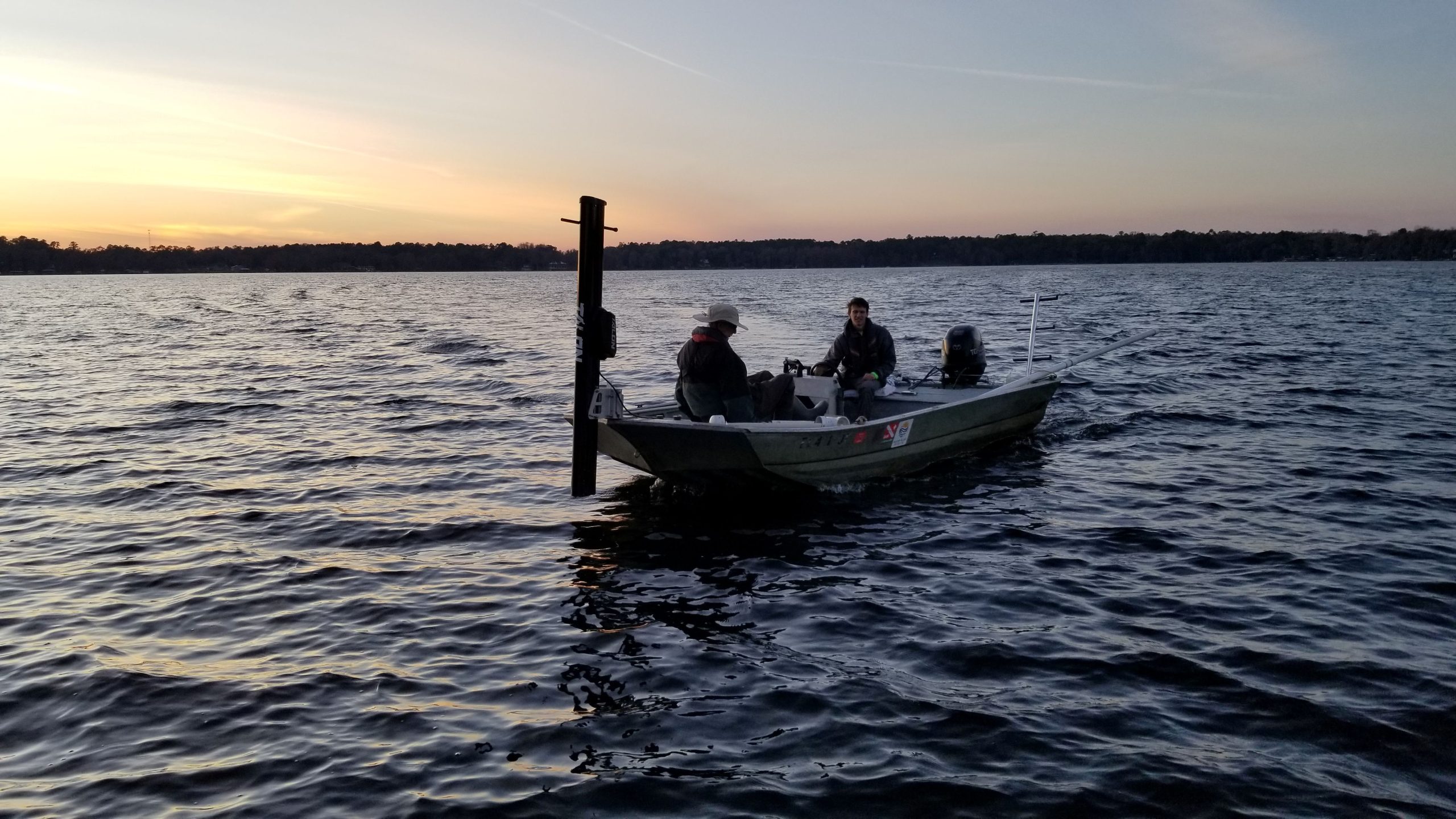 Field scientists aboard a survey boat collect environmental data on a lake during sunset.