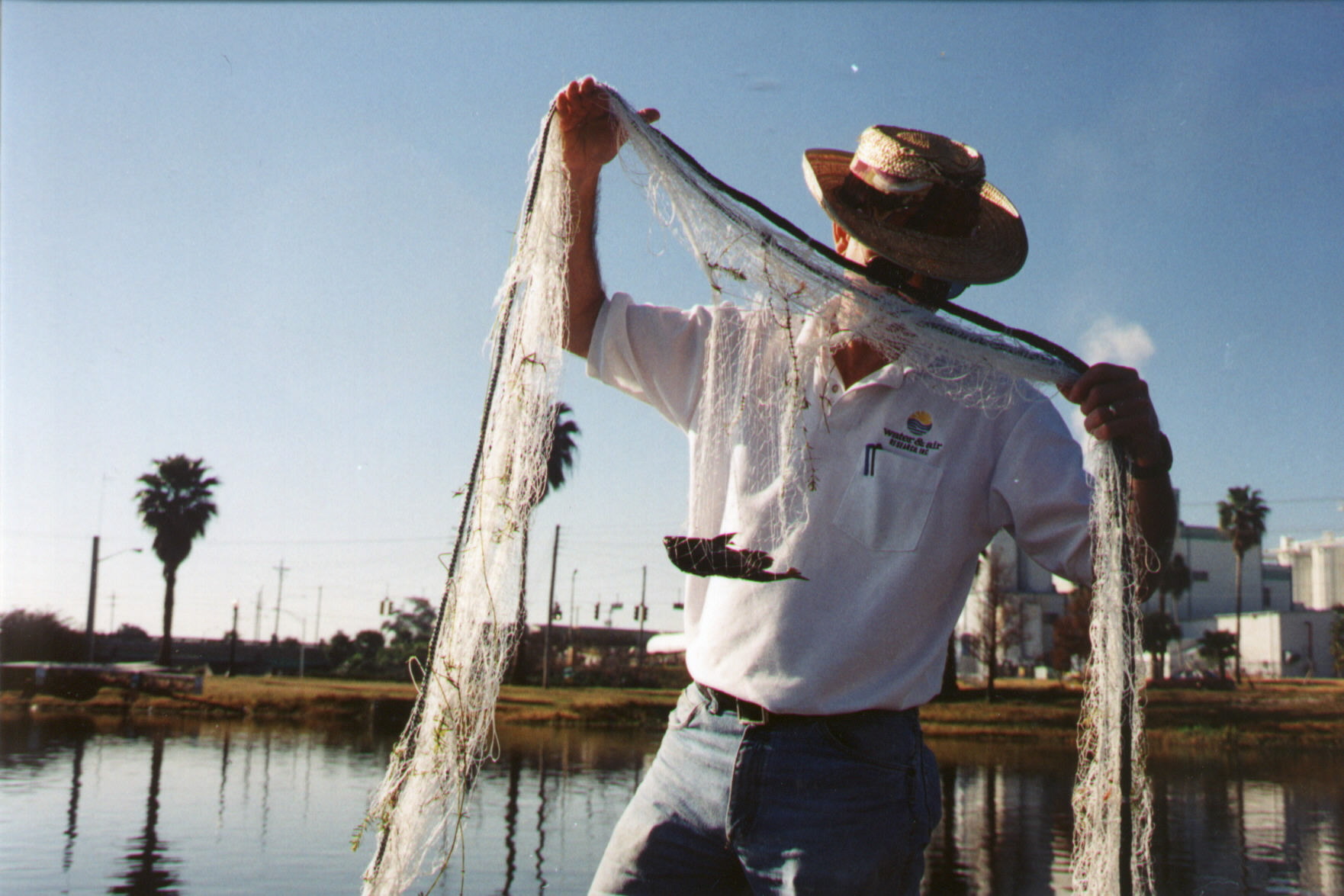A field scientist stands beside a waterway holding a seine net after sampling, with palm trees and an industrial shoreline visible in the background.